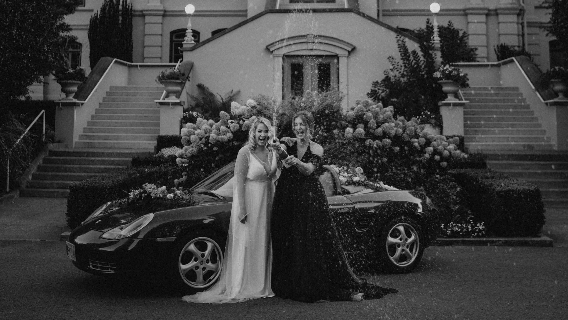 two brides in a white and black dress in front of a convertible popping champagne