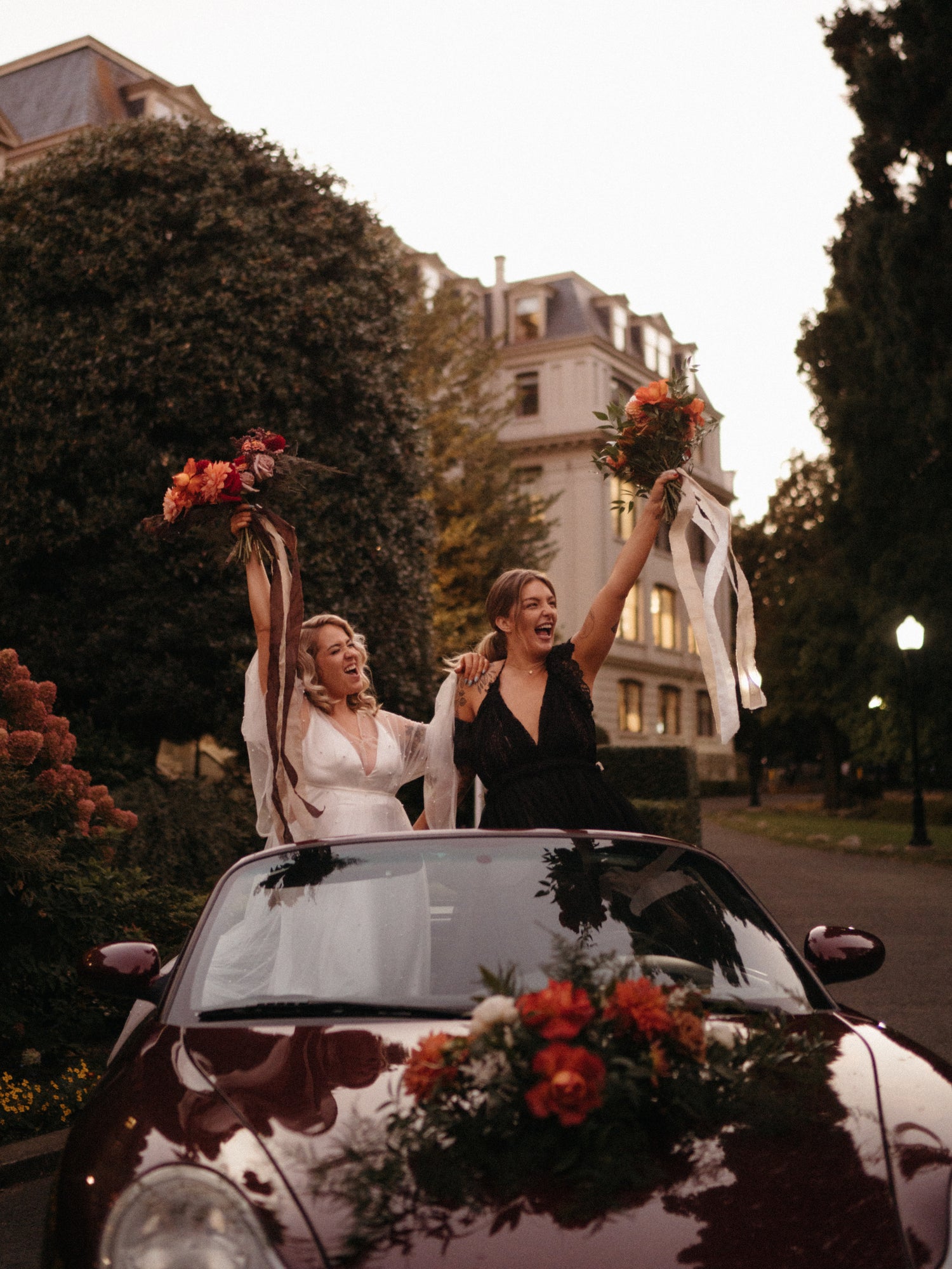 two brides cheering with their bouquets in the air standing in a convertible car