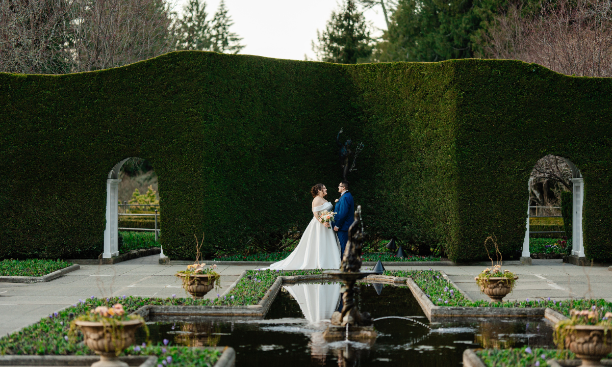 Wedding couple standing in a garden with a fountain and manicured hedges. Butchart Gardens Wedding, Victoria, BC, Photo by Jon-Mark Photography, Intended Events